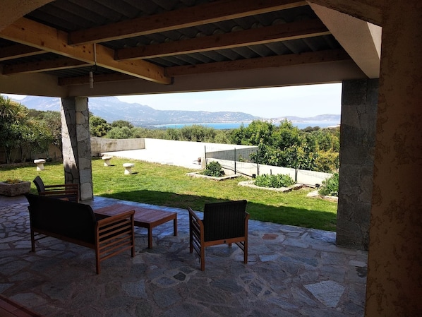 A covered patio with stone flooring and two wicker chairs, overlooking a grassy area with a view of mountains and a body of water. - MAISON 150m2 Rd C DE VILLA SUR LE GOLF DE CALVI for 4 to 8 adults clim (Lumio)