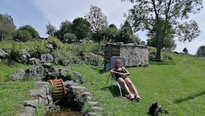 A person sitting on a chair in a grassy outdoor area with a small stream and a dog nearby. - Chalet style apartment, quiet environment and mountain sports nearby (La Bresse)