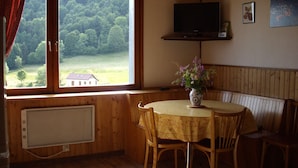 A cozy room with a wooden interior, a round table with chairs, a television on a shelf, and a large window offering a view of a forested area. - Chalet style apartment, quiet environment and mountain sports nearby (La Bresse)