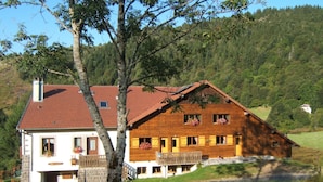 A two-story wooden house with a red-tiled roof, a stone chimney, and a balcony with flower boxes. - Chalet style apartment, quiet environment and mountain sports nearby (La Bresse)