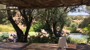 A covered outdoor area with a table, a lamp, and a view of a natural landscape including trees and a body of water. - Quiet sheepfold near Palombaggia beach with private pool. (Porto-Vecchio)