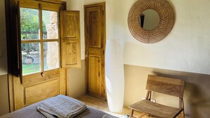A room with a wooden bed, a bench, a large mirror, and a window with a view of greenery. - Quiet sheepfold near Palombaggia beach with private pool. (Porto-Vecchio)