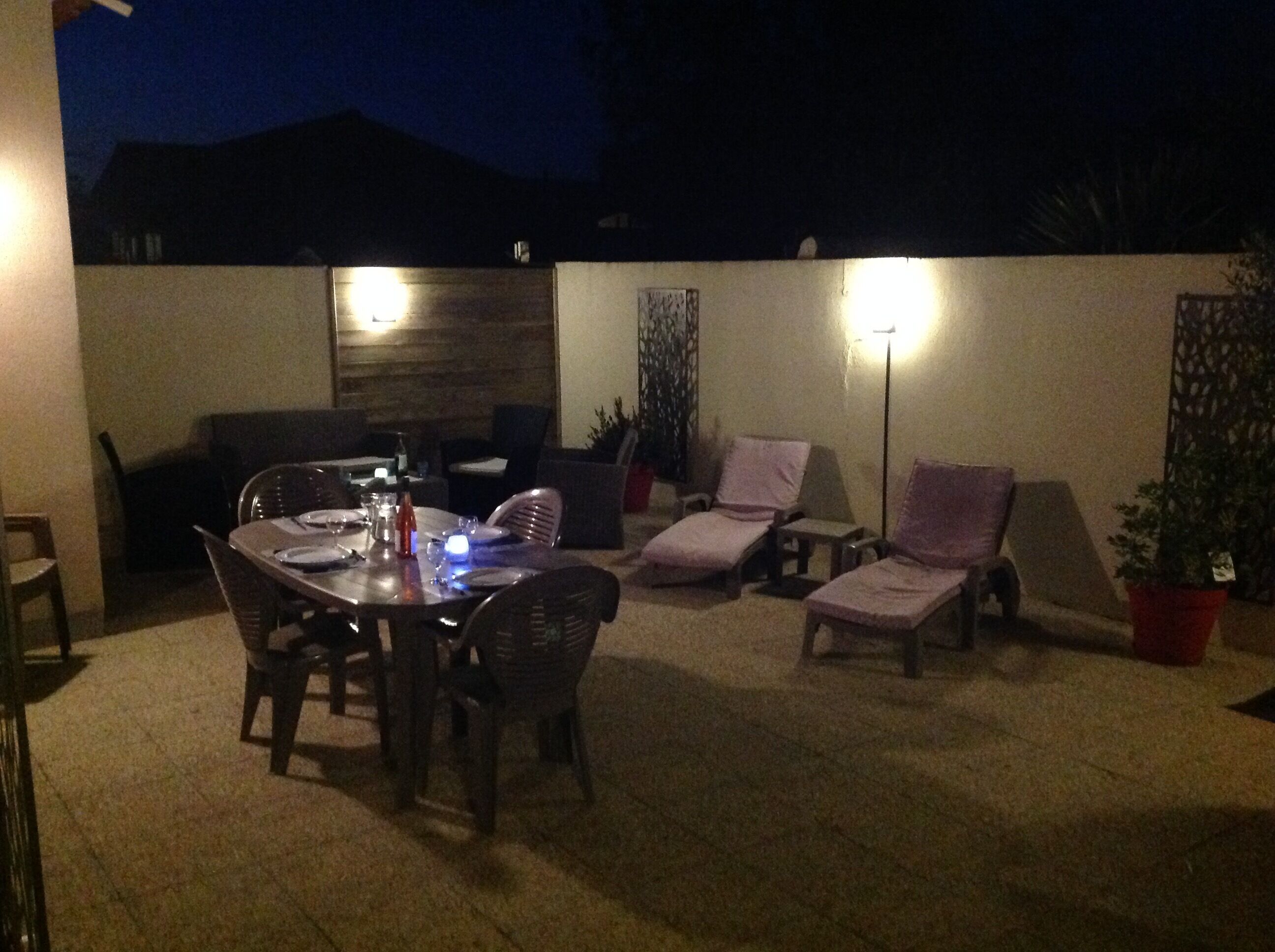 An outdoor patio area with a dining table set for four, surrounded by chairs. There are two lounge chairs with cushions. A tall floor lamp is visible, and a potted plant is placed near the lounge chairs.