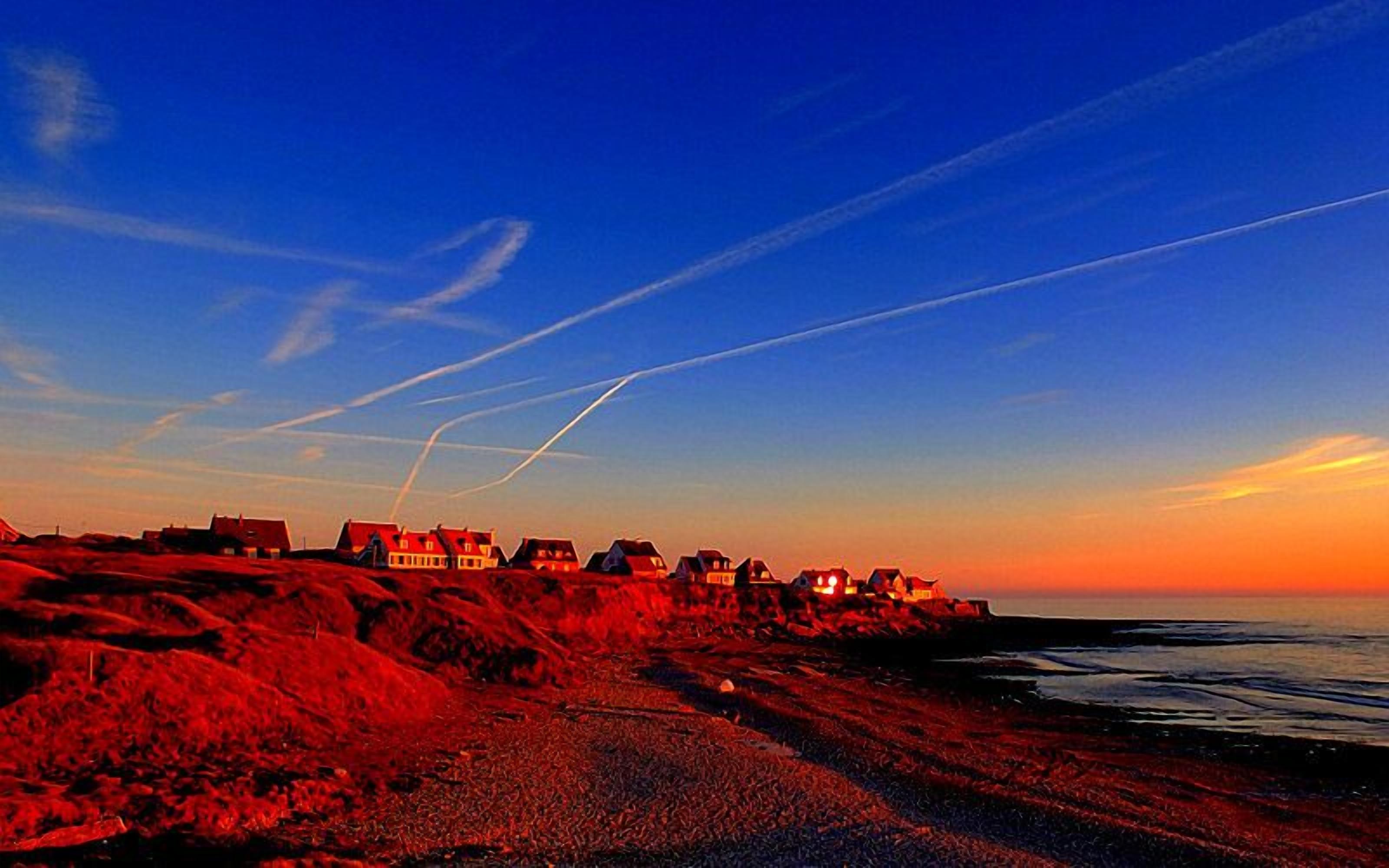 A coastal landscape with houses along the shore, a clear sky, and visible contrails.