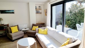 A modern living room with a white sofa, two armchairs, and a small round table. There is a bookshelf with books and decorative items, and a large window offering a view of greenery outside. - Charming new house with garden to the South of Wimereux and close to t (Wimereux)