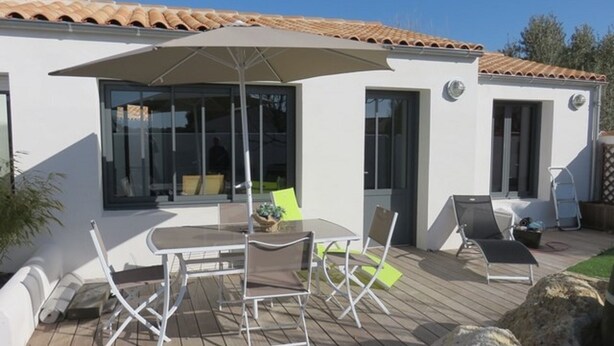 A patio with a table and chairs, a large umbrella, and a white building with a tiled roof. - Charming house close to the sea and cycle paths (Saint-Martin-de-Ré)
