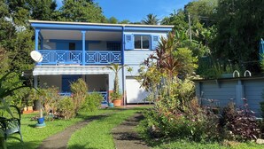Ein zweistöckiges Haus mit blauer Fassade und einem Balkon, umgeben von üppiger Vegetation und blauem Himmel.