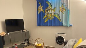 Ein Wohnzimmer mit grauem Sofa, Fernseher, Wandventilator und blauem Vorhang mit Meeresschildkrötenmuster.
