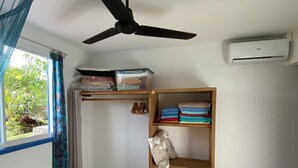 Ein Zimmer mit einem Bett, Deckenventilator, Klimaanlage, einem Fenster mit Vorhängen und einem Regal mit zusammengefalteter Kleidung.
