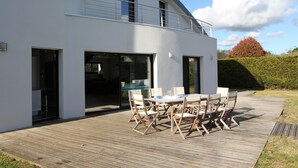 Una casa moderna de dos pisos con terraza de madera, mobiliario exterior y un gran ventanal.