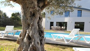 Zona de piscina con sillones blancos, una terraza de madera y un edificio moderno al fondo.