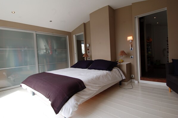 Une chambre moderne avec un grand lit, une porte-fenêtre coulissante et une petite table de chevet avec une lampe.