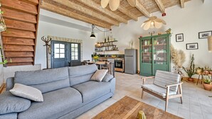 A cozy living room with a sofa, a wooden coffee table, and a patterned armchair. A kitchen with a dining area is visible in the background. - La Maison des Vendangeurs***, Gîte with swimming pool and bicycles  (Beaucaire)
