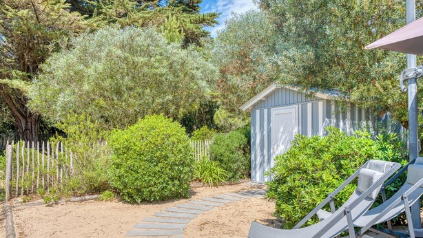 A wooden deck with a sandy path leading to a small shed, surrounded by dense greenery and a wooden fence.