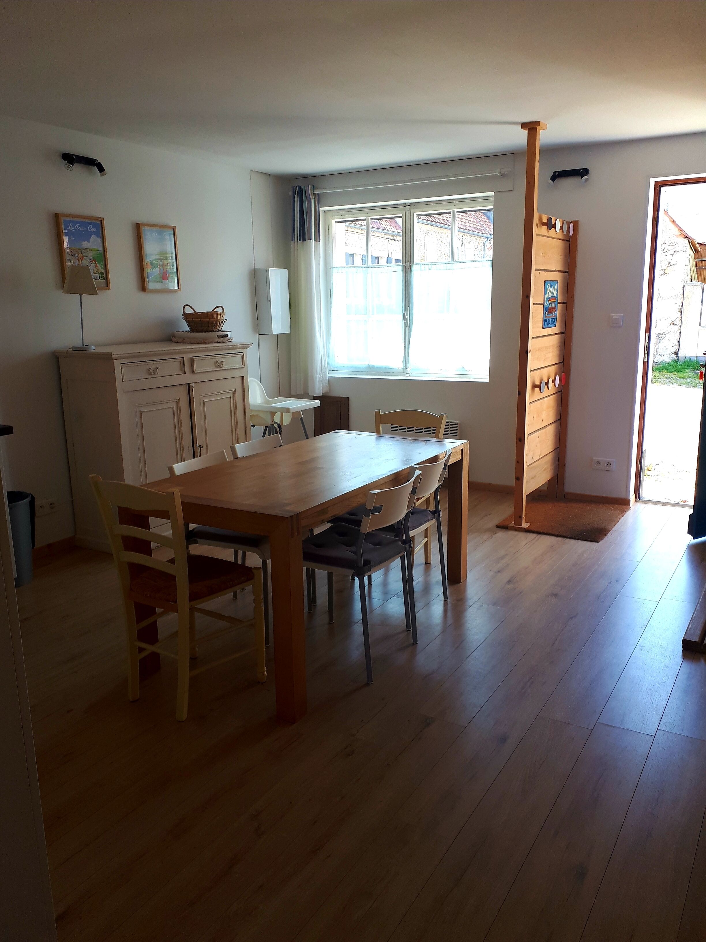 Una habitación con una mesa de comedor de madera, sillas, un armario blanco, una ventana con cortinas y una puerta que da al exterior.