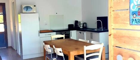 Een compacte keuken met een houten tafel en stoelen, een magnetron op een witte koelkast en een koffieapparaat.