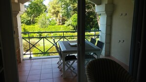 En balkon med et bord og stole, udsigt over en velplejet have, og en dør der fører ind.