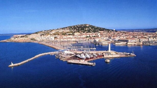 An aerial view of a coastal town with a marina, a lighthouse, and a hill with buildings.