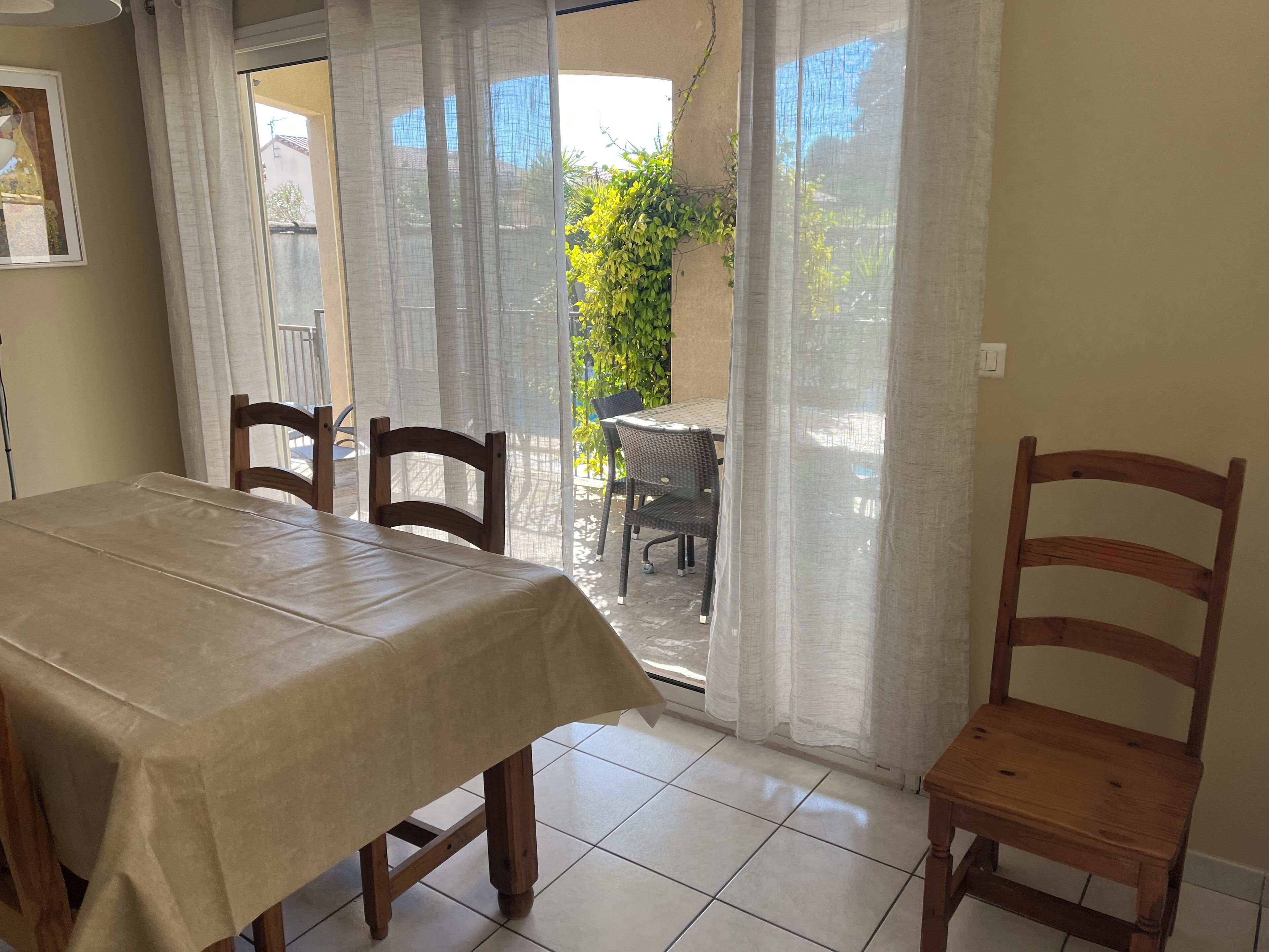 A dining area with a table covered by a beige tablecloth, wooden chairs, and a view through a glass door to an outdoor patio with a chair and greenery.