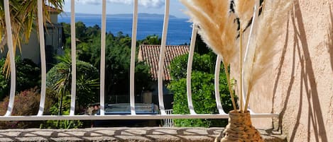 A balcony with a woven vase containing pampas grass, a railing, and a view of the sea and buildings.