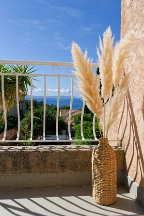 Un balcon orné d’un vase en osier contenant des plumes d’herbe des pampas, une balustrade et une vue sur la mer et les bâtiments.