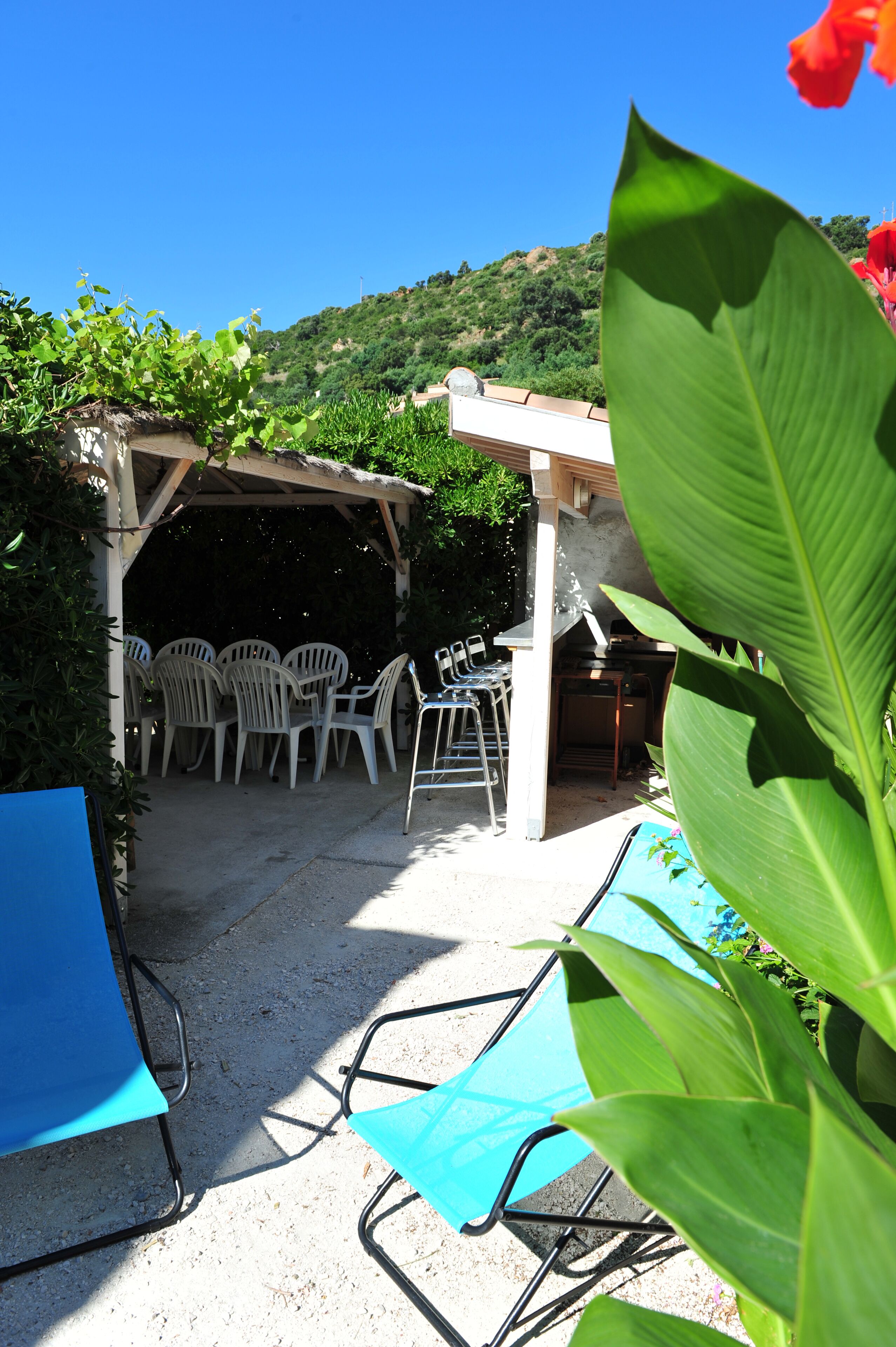 Eine Terrasse im Freien mit weißen Plastikstühlen und einem Tisch, umgeben von viel Grün und blauem Himmel.