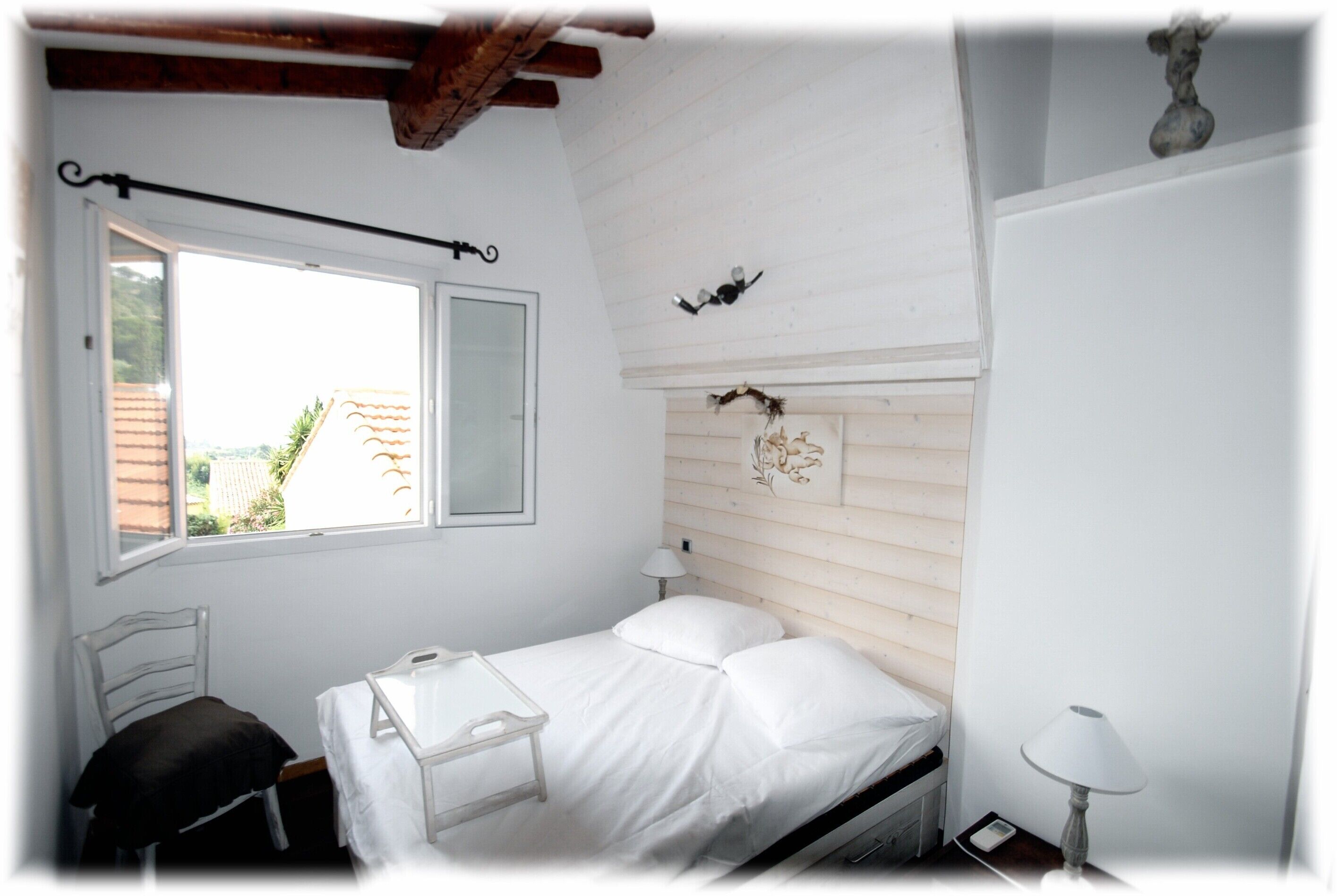 A small, white room with a bed, a window, a bedside table, and a lamp.