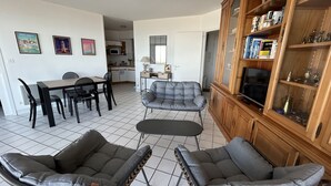 Een woonkamer met een eettafel, twee grijze fauteuils, een bank en een houten kast met glazen deuren.