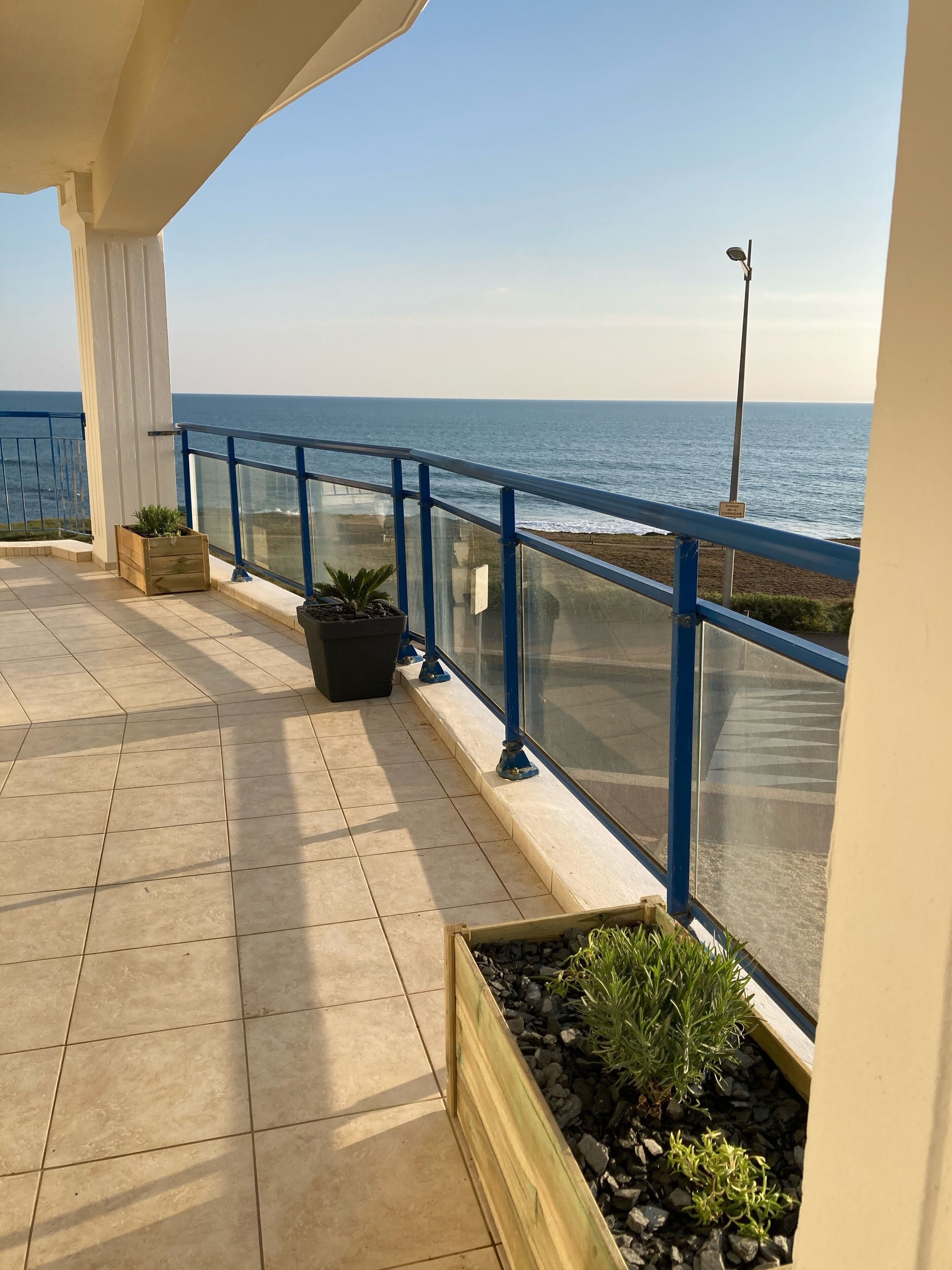 A balcony with a view of the sea, featuring tiled flooring, potted plants, and a blue railing.