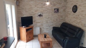 A living room with a TV on a wooden stand, a black leather sofa, a small wooden table, and a stone wall. - Recently built house, nice setting, 500 m from sea, at heart of pedestrian hill (Tréveneuc)