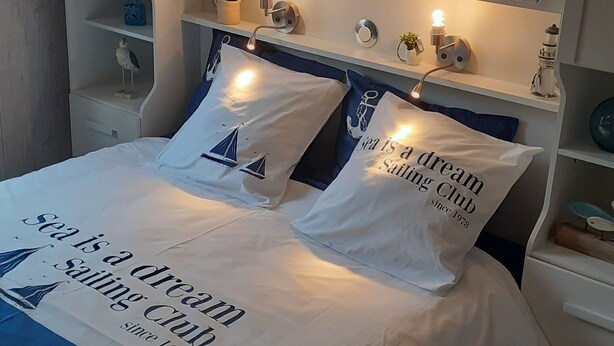 A neatly made bed with a blue and white striped bedspread and pillows, a white headboard, and bedside tables with small decorative items. - Recently built house, nice setting, 500 m from sea, at heart of pedestrian hill (Tréveneuc)