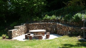 A stone patio with a round table and benches, surrounded by a stone wall and lush greenery. - Recently built house, nice setting, 500 m from sea, at heart of pedestrian hill (Tréveneuc)