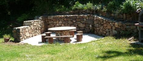 A stone patio with a round table and benches, surrounded by a stone wall and lush greenery.