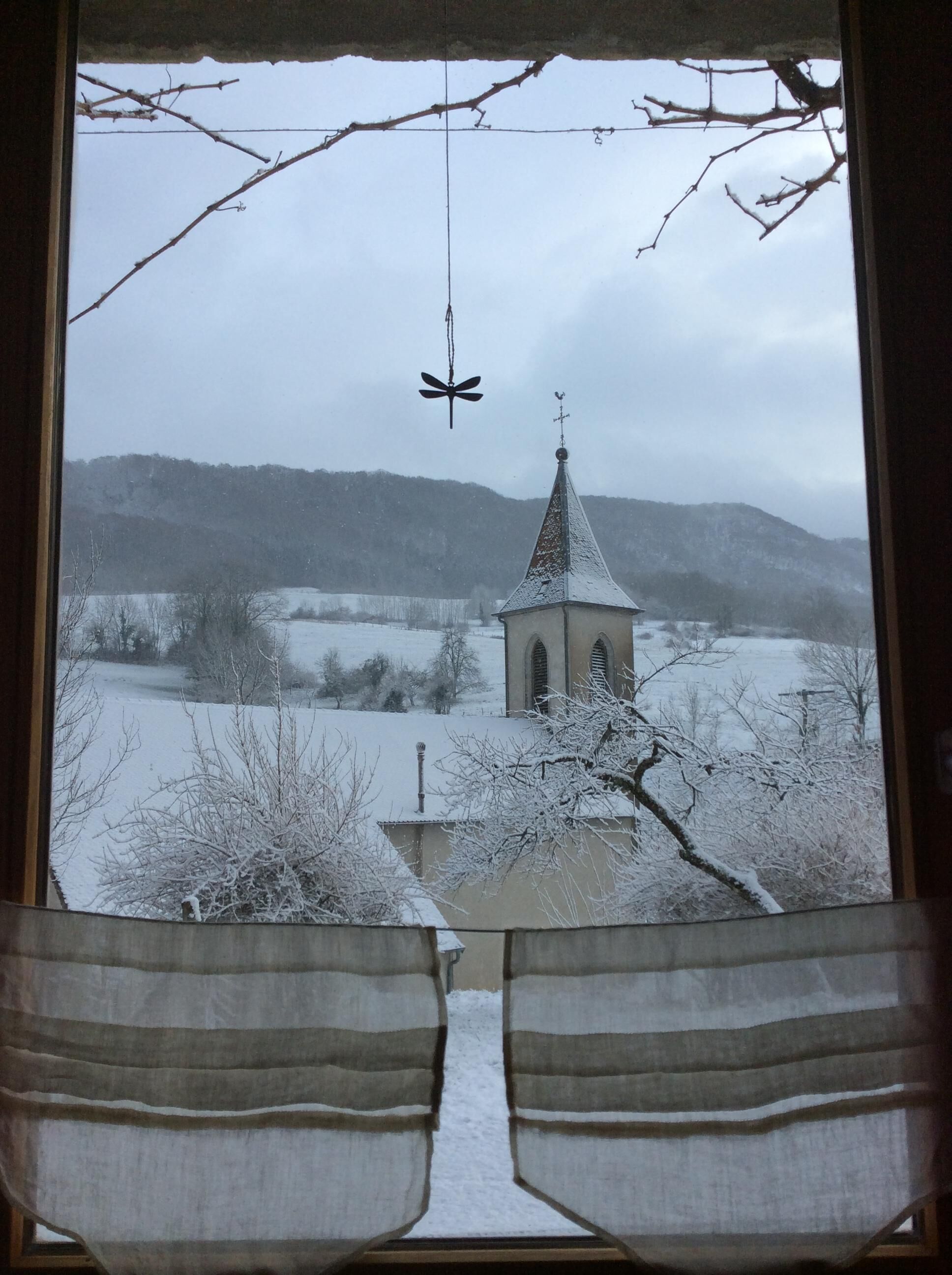Une vue à travers une fenêtre avec des rideaux, donnant sur un paysage enneigé, une église avec un clocher et des arbres sans feuilles.