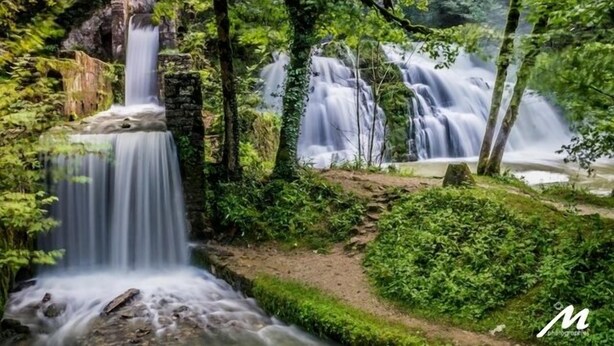 A waterfall in a lush forest setting with a stone structure and a clear path leading to the water.