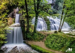 A waterfall in a lush forest setting with a stone structure and a clear path leading to the water. - Cottage character in quiet village on the slopes of Mount Poupet (Ivrey)