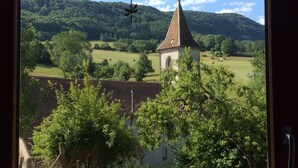 A view through a window with curtains, featuring a church, trees, and a hill in the background.