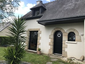 Une maison avec une façade en pierre, une porte noire et un petit jardin agrémenté d’un palmier.