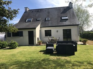 Une maison à deux étages avec un toit en pente, une terrasse dotée de mobilier d’extérieur et un petit jardin.