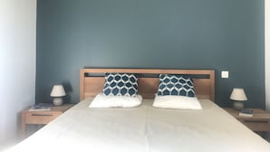 Un lit bien fait, agrémenté de deux coussins à motifs, d’une tête de lit en bois et de deux tables de chevet sur lesquelles reposent des lampes et des livres.