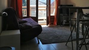Quarto com cama, escrivaninha e vista para montanhas através de janelas amplas.