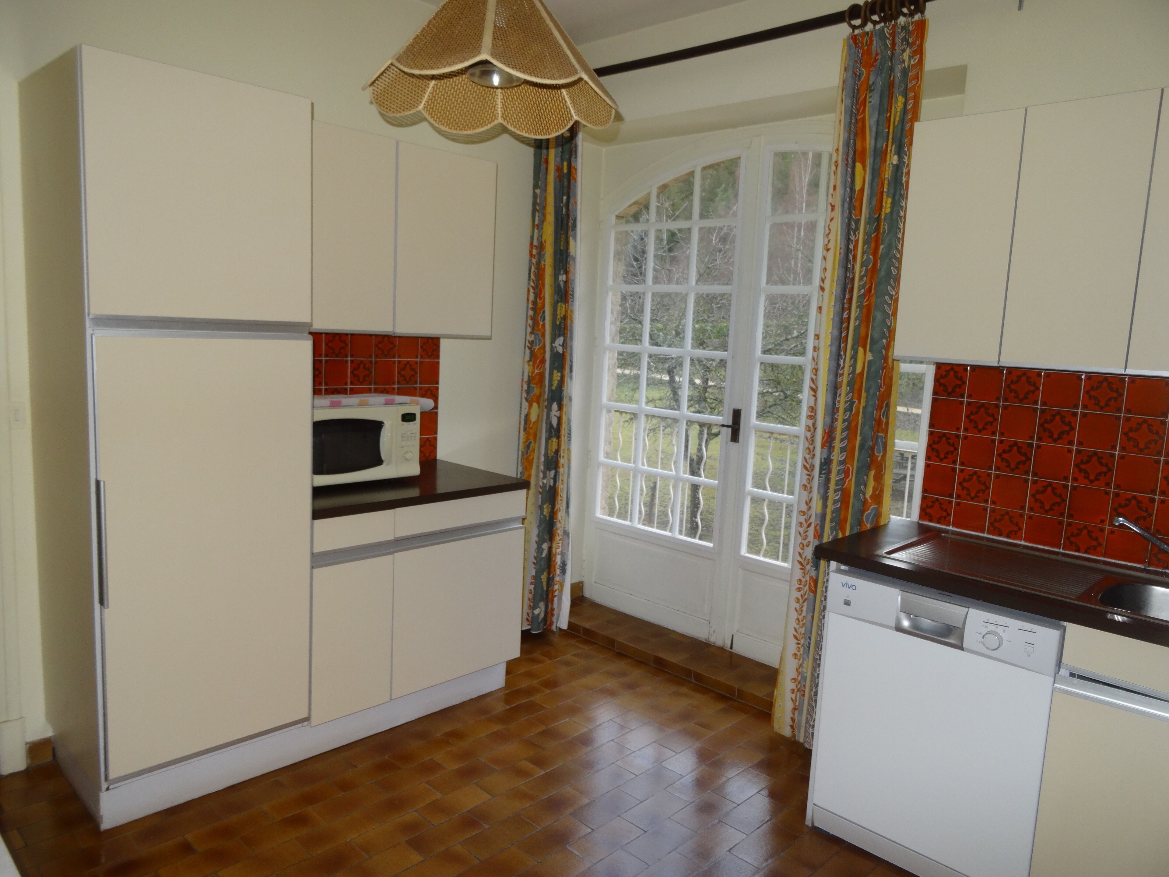Una cocina con armarios blancos, microondas, lavavajillas y una ventana con cortinas.