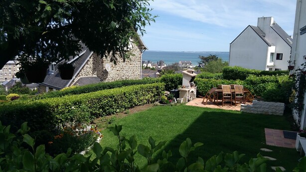 Ein gepflegter Garten mit einem Steinhaus, einer Holzterrasse mit Stühlen und Blick auf das Meer.