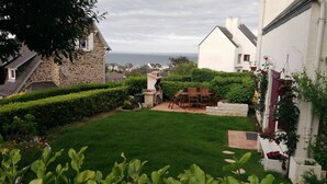 Ein gepflegter Garten mit einem Steingebäude, einem Holztisch und -stühlen und Blick auf das Meer.