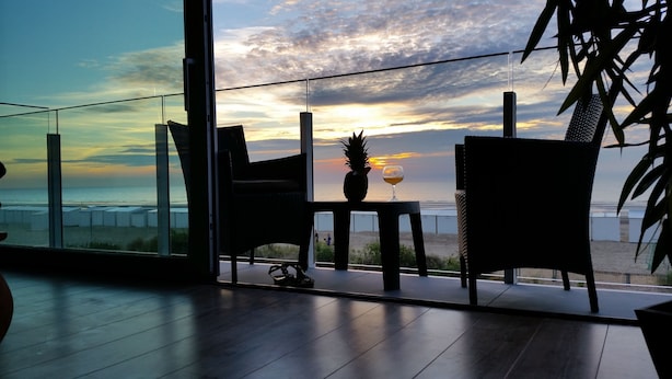 A balcony with a view of the sunset over the ocean, two chairs, a table with a pineapple and a glass of wine, and a plant.