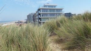 A modern multi-story building with a glass facade, located near a beach with tall grass in the foreground.