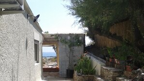 Un étroit sentier mène à un bâtiment doté d’un mur en pierre et offrant une vue sur la mer. On y trouve des plantes en pot et une structure surmontée d’un toit de chaume.