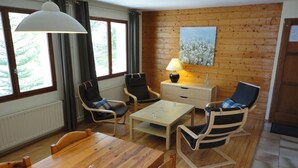 A cozy room with a wooden floor, a wooden dining table, two armchairs, a sofa, a lamp, and a painting on the wall.