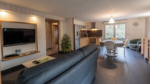 Ein modernes Wohnzimmer mit einem großen Flachbildfernseher, einem grauen Sofa, einem Essbereich mit Tisch und Stühlen sowie einer Küche mit Herd und Kühlschrank.