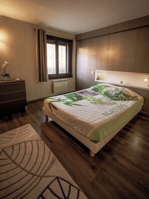 A bedroom with a bed, a dresser, a window with curtains, a radiator, and a bedside table with a lamp. - GÎTE 3 STARS, BESSE & ST-ANASTAISE, Theme: SKIING, HIKING, VISITS, FISHING (Besse-Et-Saint-Anastaise)